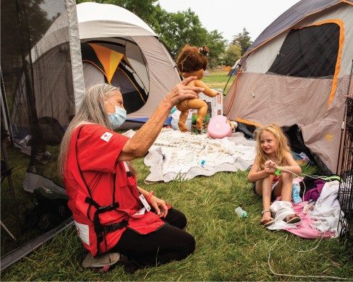 red cross volunteer blowing bubbles and playing with a young girl outside a temporary shelter